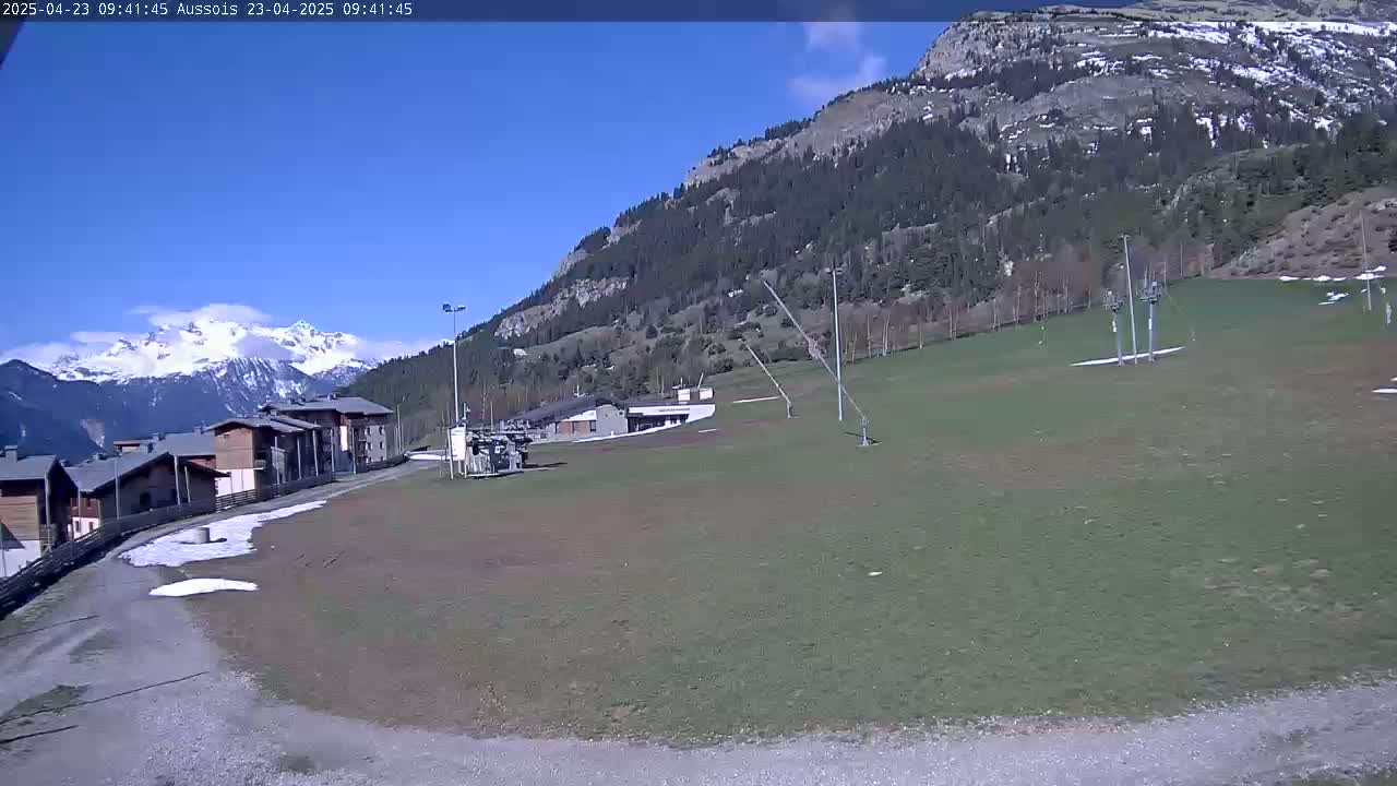 A mostly sunny day reveals a grassy slope with a ski lift, a cluster of buildings, and snow-capped mountains in the distance.