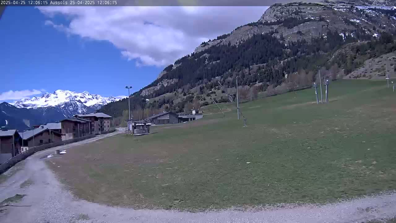 A partly cloudy day shows a grassy field with buildings and a road leading to snow-capped mountains in the background.