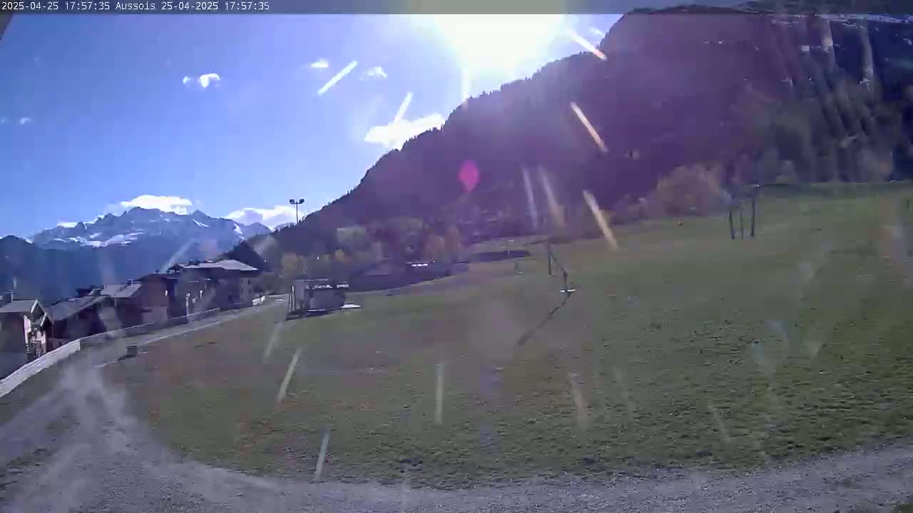 A sunny day with light snow or rain shows a view of a grassy field, some buildings, and mountains in the distance.