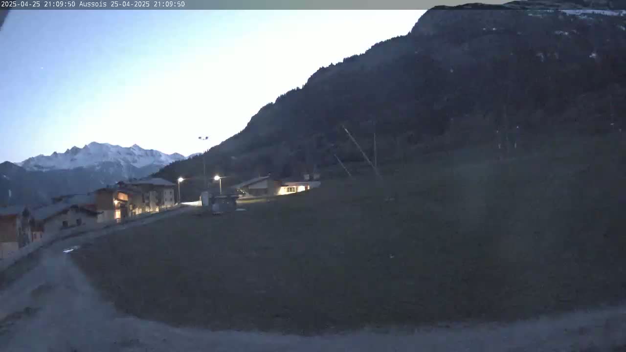 A village nestled at the base of a large, dark mountain is illuminated by twilight, with snow-capped mountains visible in the distance.