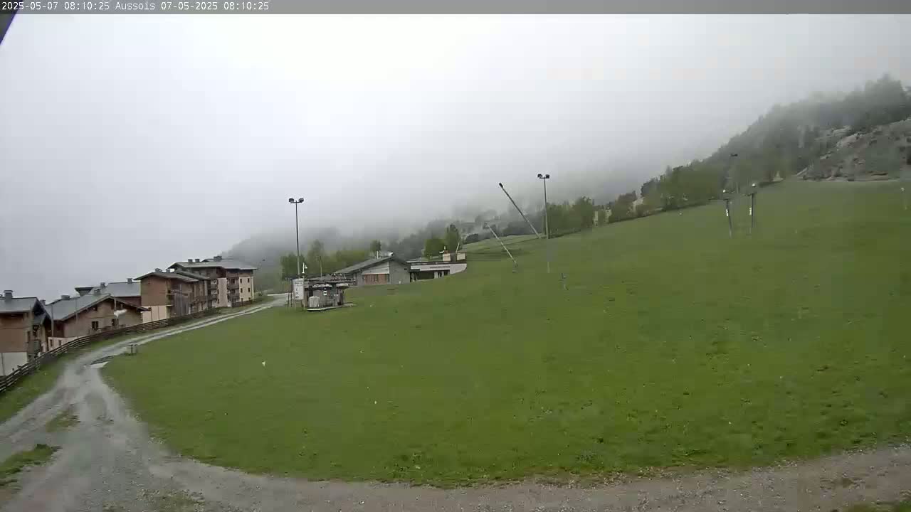 A foggy mountainside view shows a grassy area with buildings and ski lift equipment in the foreground.