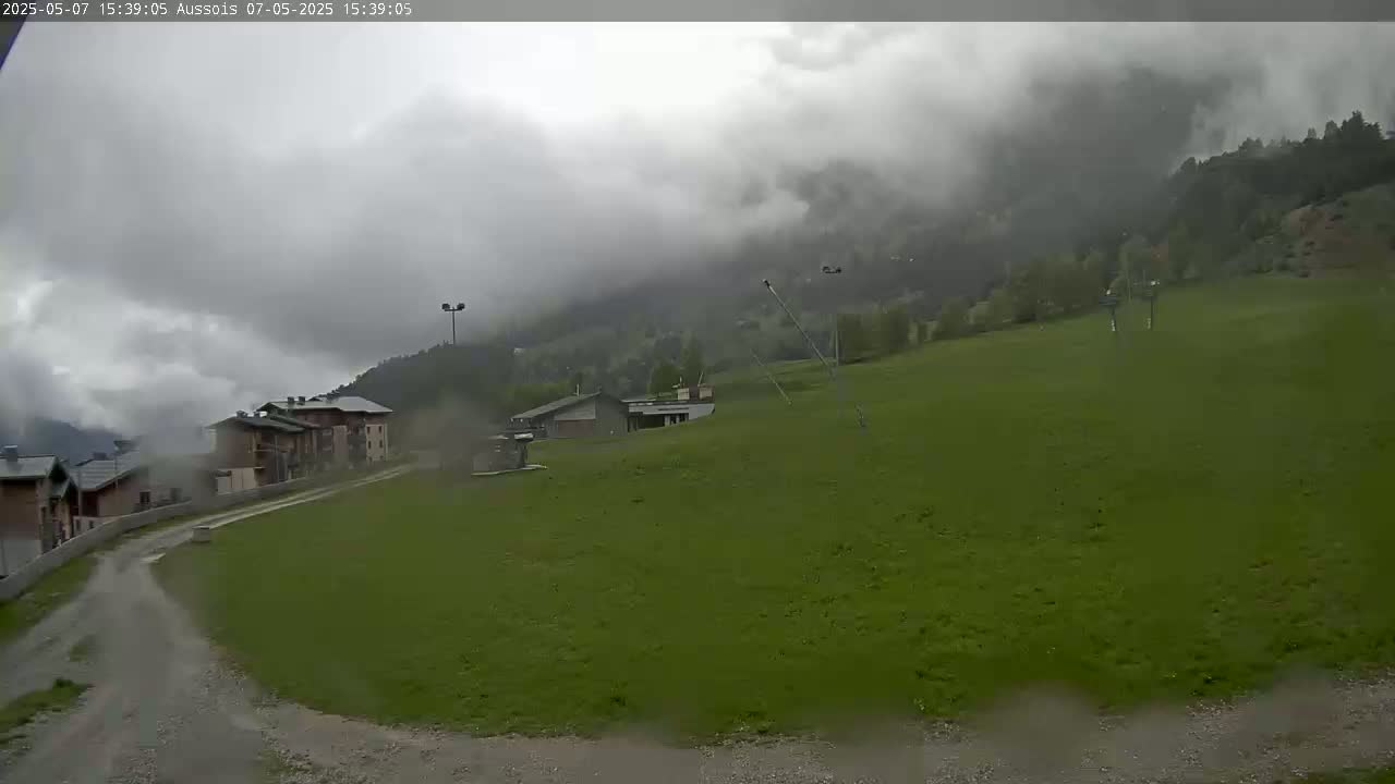A grassy field with low buildings and a mountainside shrouded in heavy clouds under overcast skies.