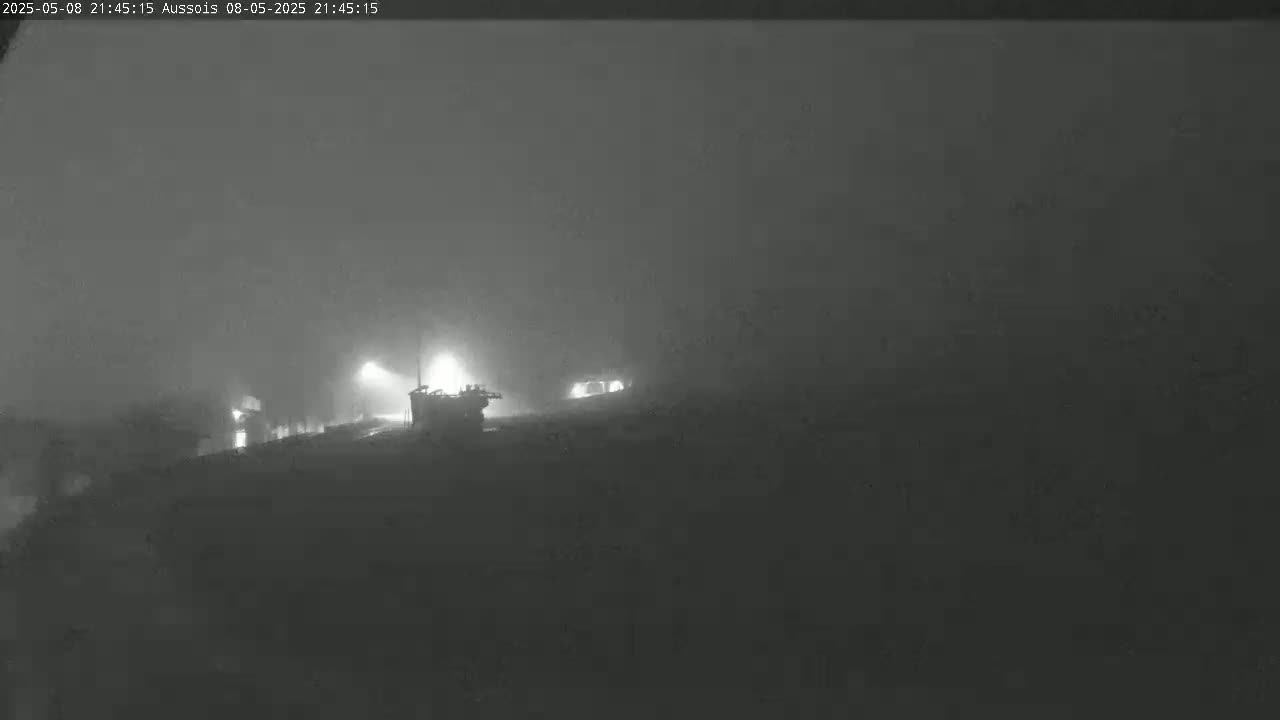 A dimly lit, foggy landscape at night shows some structures with lights in the distance.