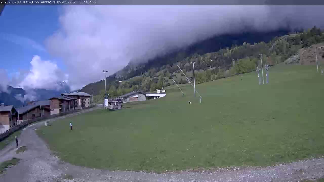 A partly cloudy day reveals a grassy field with a few buildings and a mountainside shrouded in fog.