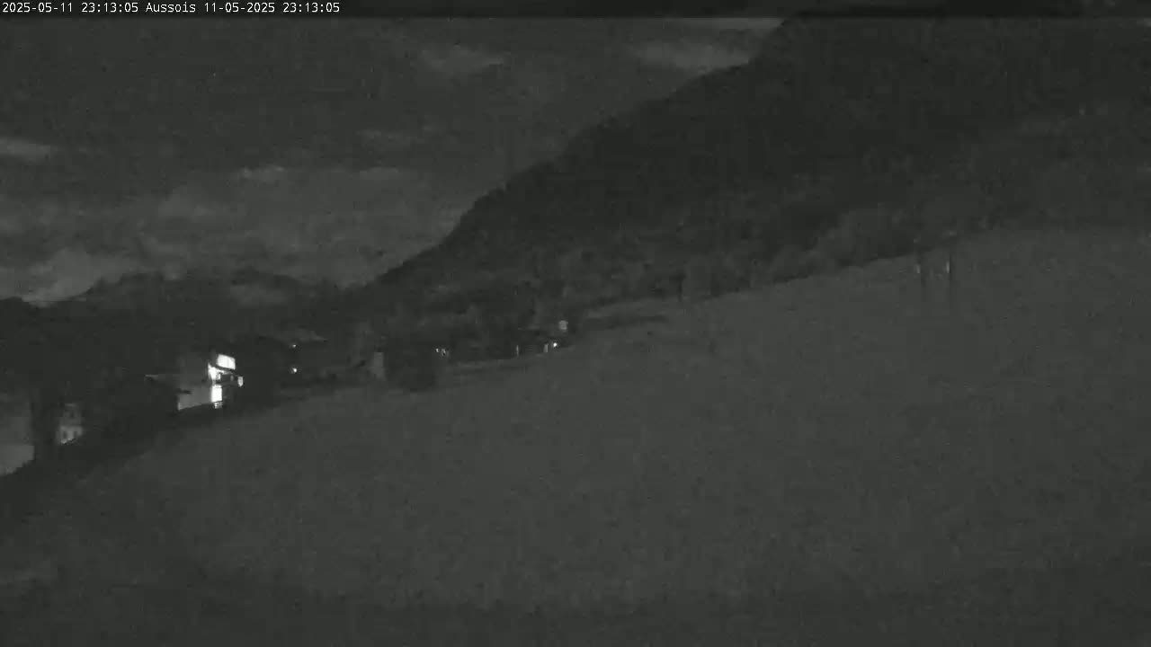 A nighttime, low-light view shows a dark hillside with a few faint lights visible in the distance under a partly cloudy sky.