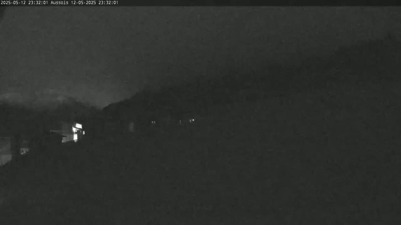 A nighttime, low-light view shows a few dimly lit buildings and a dark, possibly overcast sky.