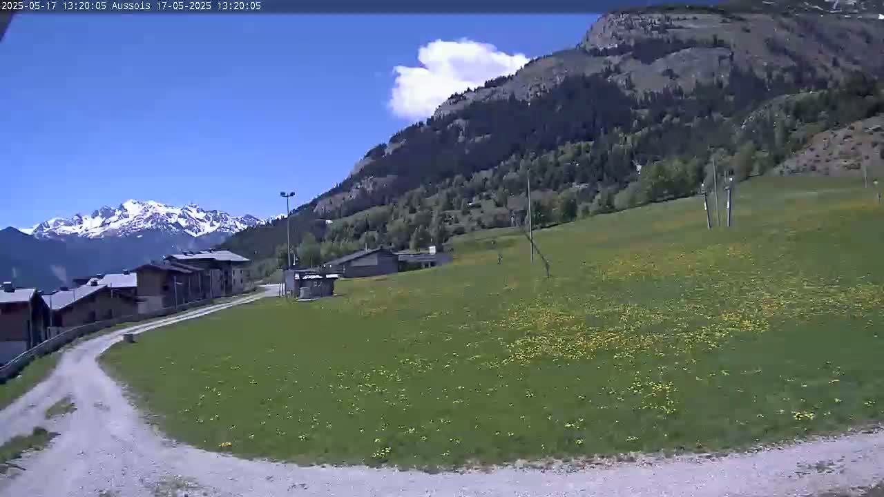 A gravel road leads to a grassy field dotted with yellow flowers, nestled between a village of low buildings and a mountainside under a partly cloudy blue sky.