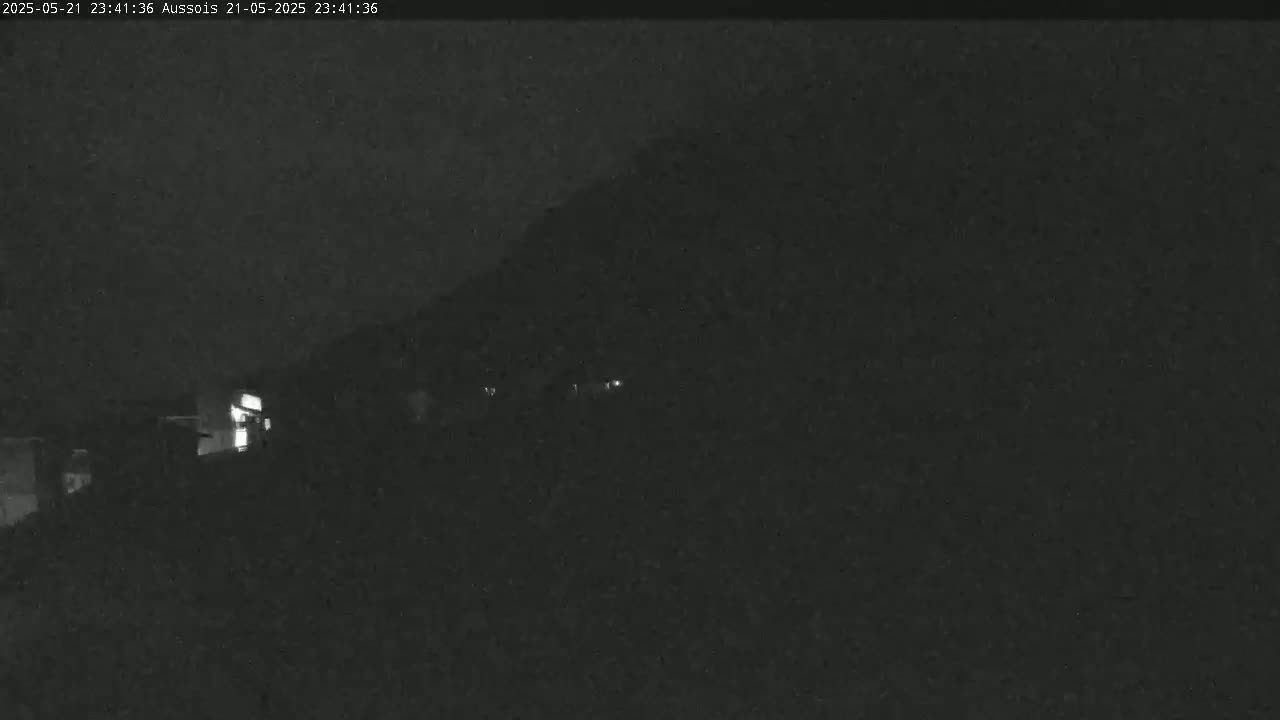 A nighttime, low-light view shows a dark hill or mountain in the background with faintly illuminated structures in the foreground.