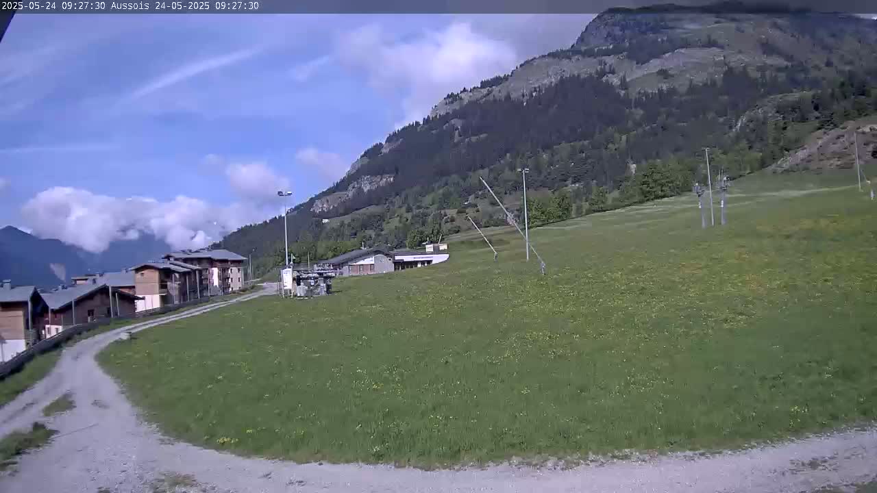 A partly cloudy day reveals a grassy field, a road, several buildings, and a mountainside covered in trees.