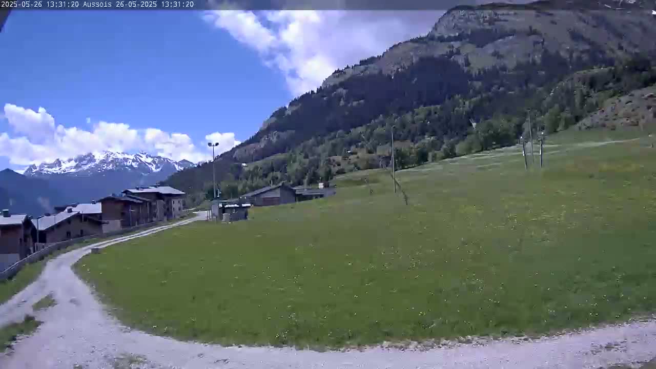 A partly cloudy day reveals a mountainside village with a grassy field and a dirt road leading to snow-capped mountains in the distance.