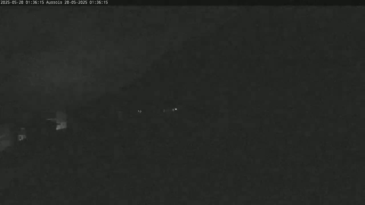 A nighttime, dark image shows indistinct shapes of buildings and a few faint lights in the distance under a cloudy sky.