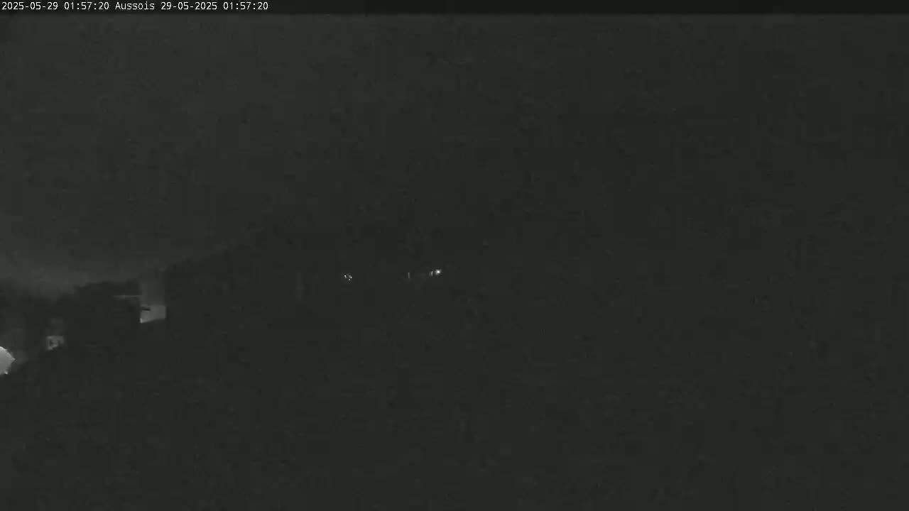 A nighttime, low-light image shows indistinct dark shapes, possibly buildings, with a few faint lights visible in the distance.