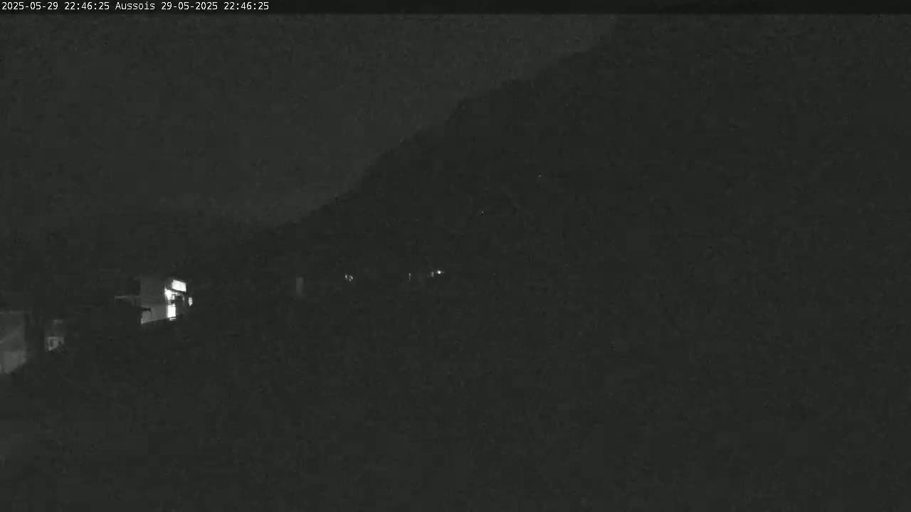 A nighttime, low-light view of a dark hillside with faintly visible structures and a few illuminated spots.