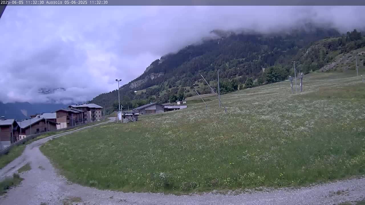 A cloudy day reveals a mountainside village with a grassy field in the foreground, featuring a dirt road leading toward buildings and a ski lift.