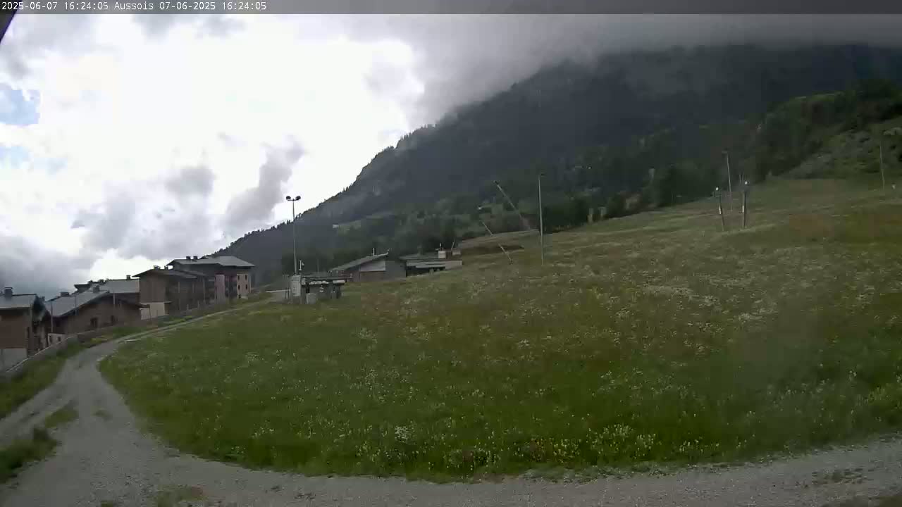 A cloudy day reveals a grassy field leading to buildings and a mountain partially obscured by clouds.