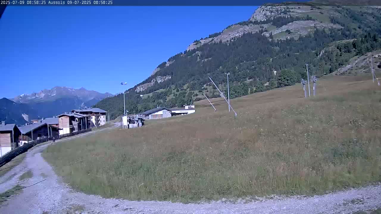 A gravel path leads to a cluster of buildings nestled in a grassy field beneath a mountainside on a clear, sunny day.