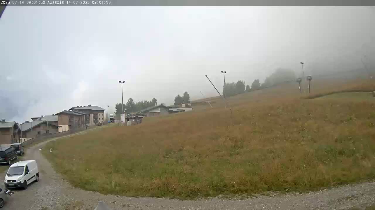 A foggy, overcast day reveals a grassy hillside with buildings and a few vehicles parked near a roadway.