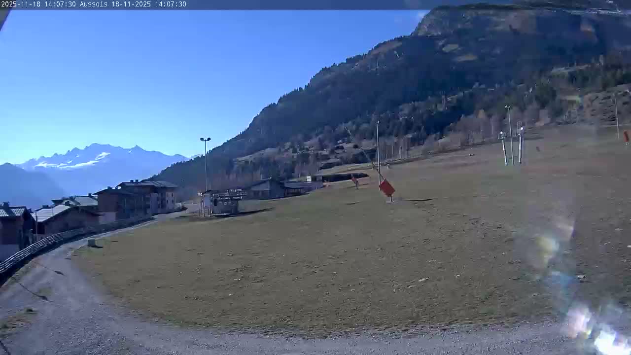 A person walks a dog on a dirt road near a village nestled at the base of a mountain under a mostly clear, sunny sky.