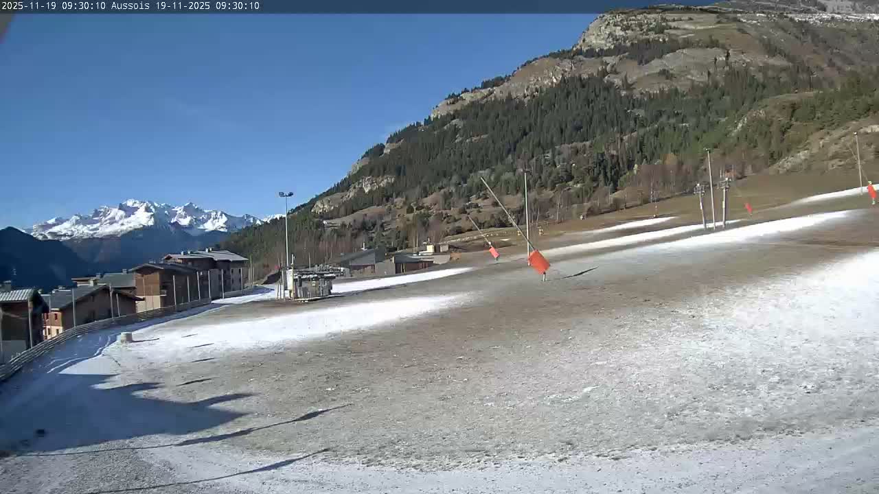 On a clear, sunny day, a partially snow-covered ski slope with buildings and prominent ski lift infrastructure stretches towards evergreen-dotted mountains, with distant snow-capped peaks under a brilliant blue sky.