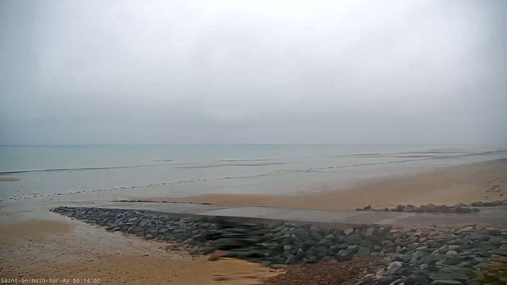 A rocky embankment leads to a flat, sandy beach extending to a calm, gray sea under an overcast sky.