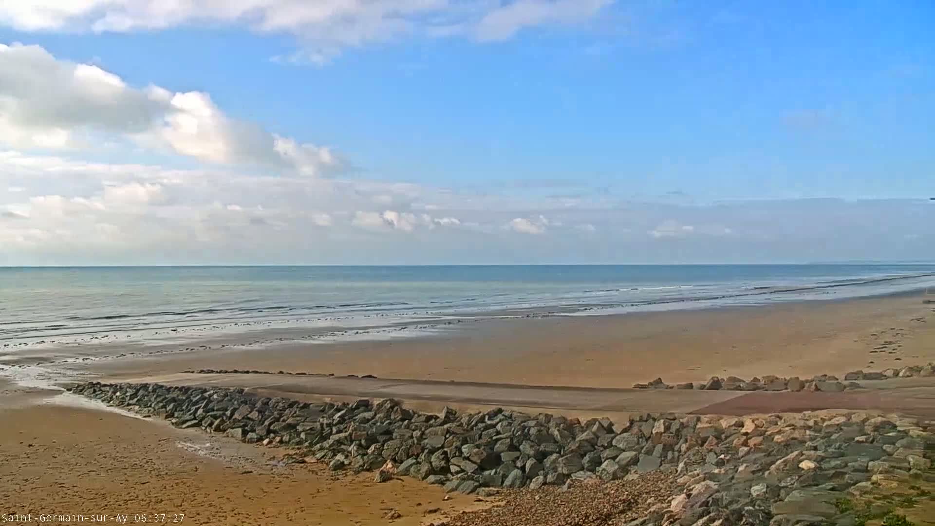 A wide sandy beach stretches to a calm ocean under a mostly sunny sky with scattered cumulus clouds.