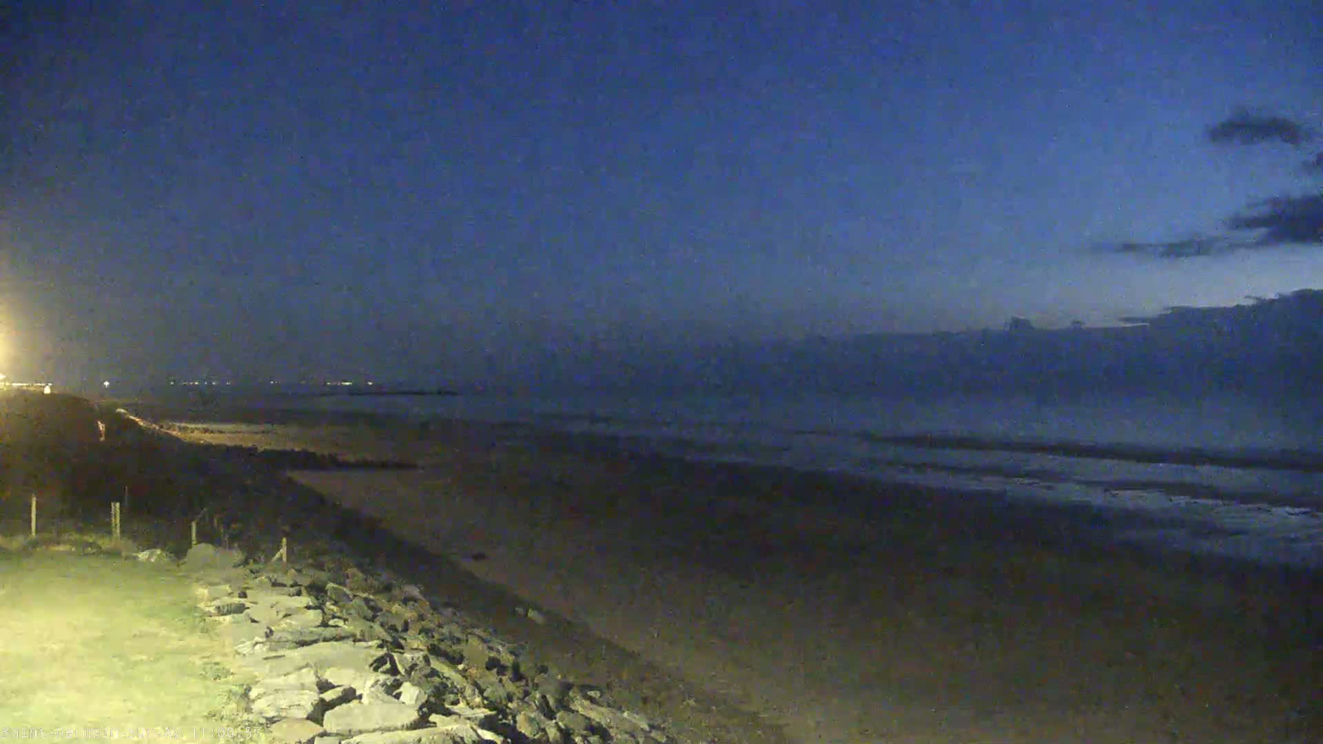 A dark beach at night under a mostly clear, twilight sky is seen from a rocky embankment.