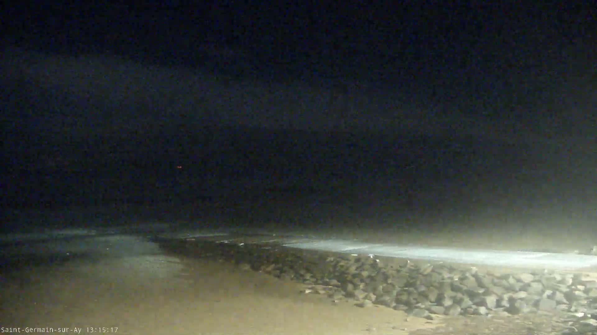 A dark, nighttime beach scene shows a rocky wall separating the sand from the water under a hazy, dark sky.