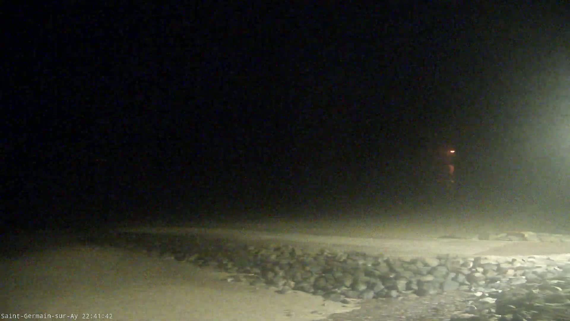 A dimly lit beach at night is shown, with a rocky wall in the foreground and a hazy atmosphere.