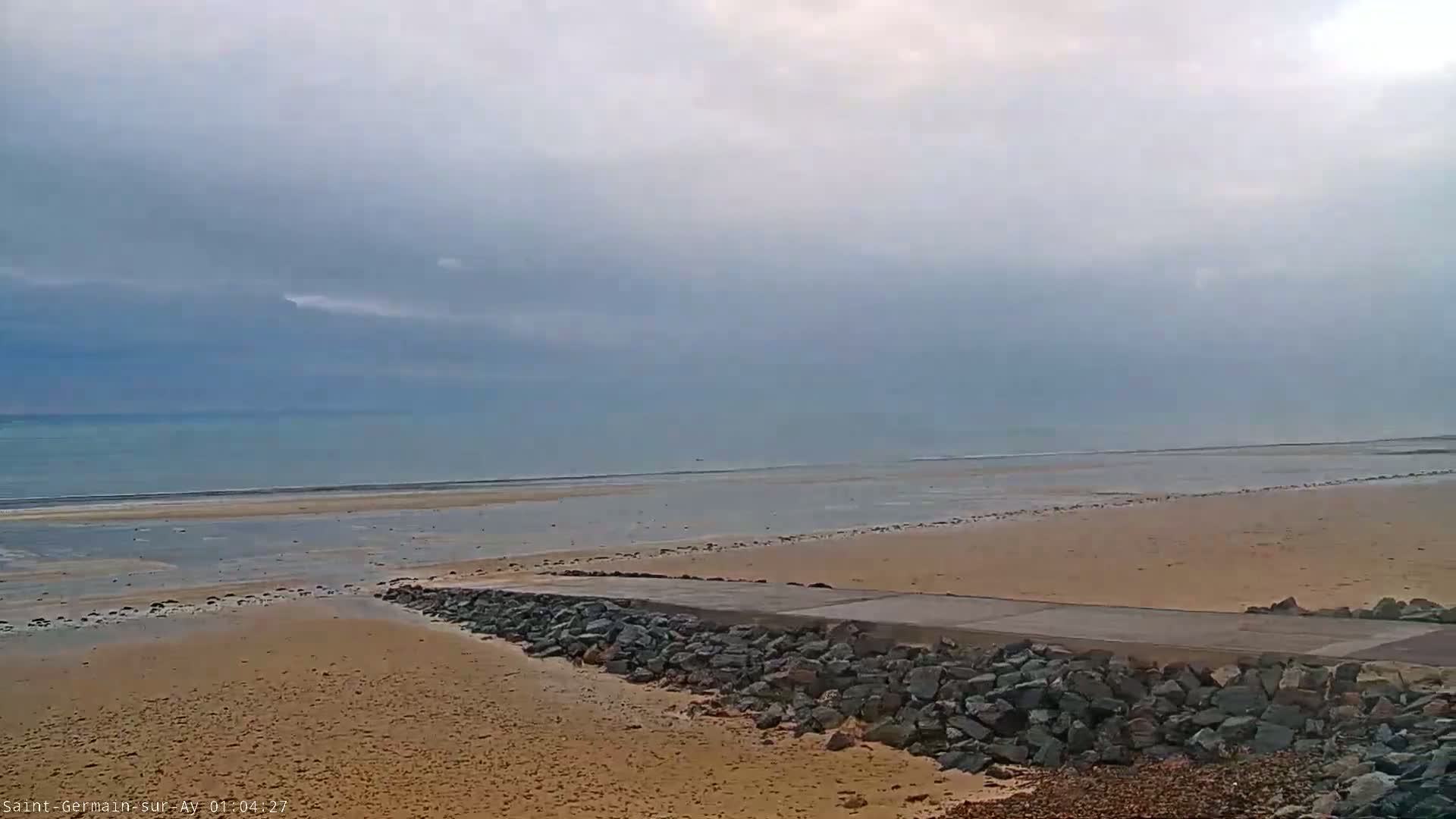A flat, sandy beach stretches to a calm, light blue ocean under an overcast sky.