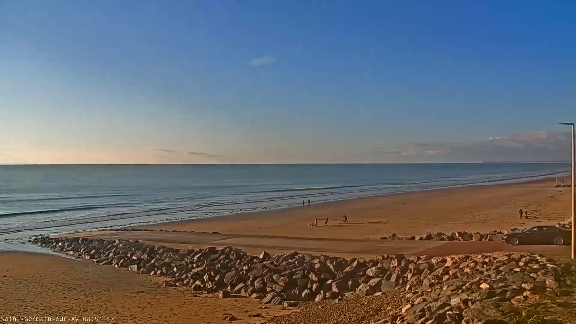 A sandy beach stretches along a calm ocean under a clear, bright blue sky, with a few people visible on the sand and a car parked near a rock wall.