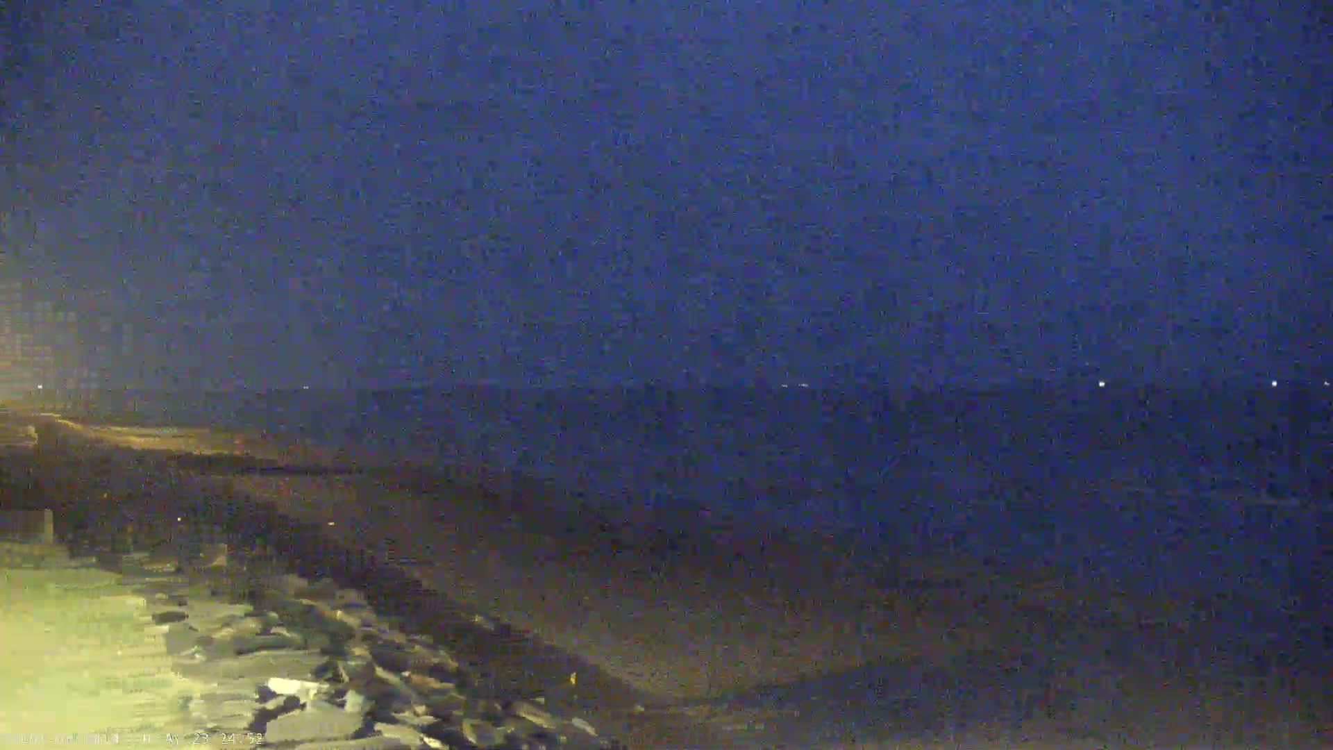 A dark, nighttime view shows a body of water under a twilight sky, with a shoreline featuring a rocky embankment in the foreground.