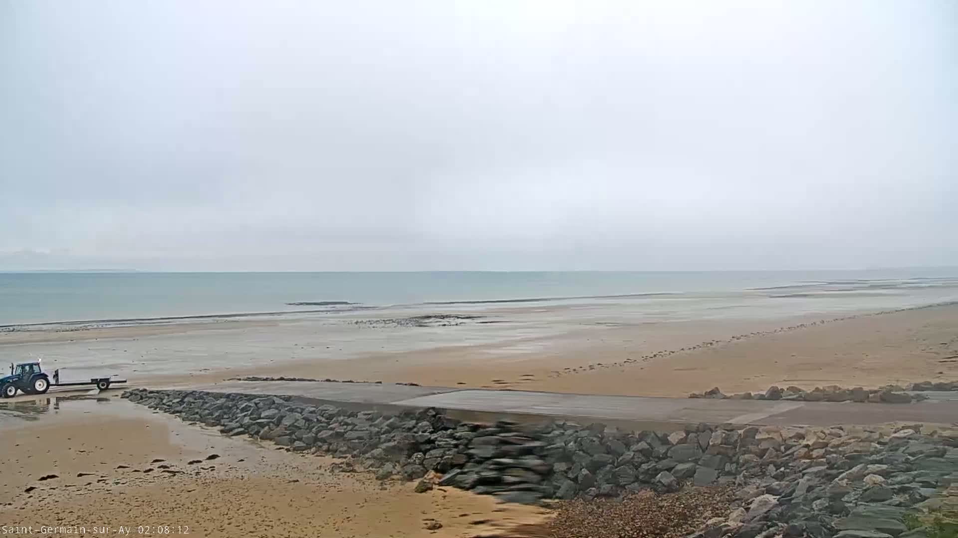 A blue tractor and trailer are parked on a sandy beach near a rocky embankment, with an overcast sky and calm sea in the background.