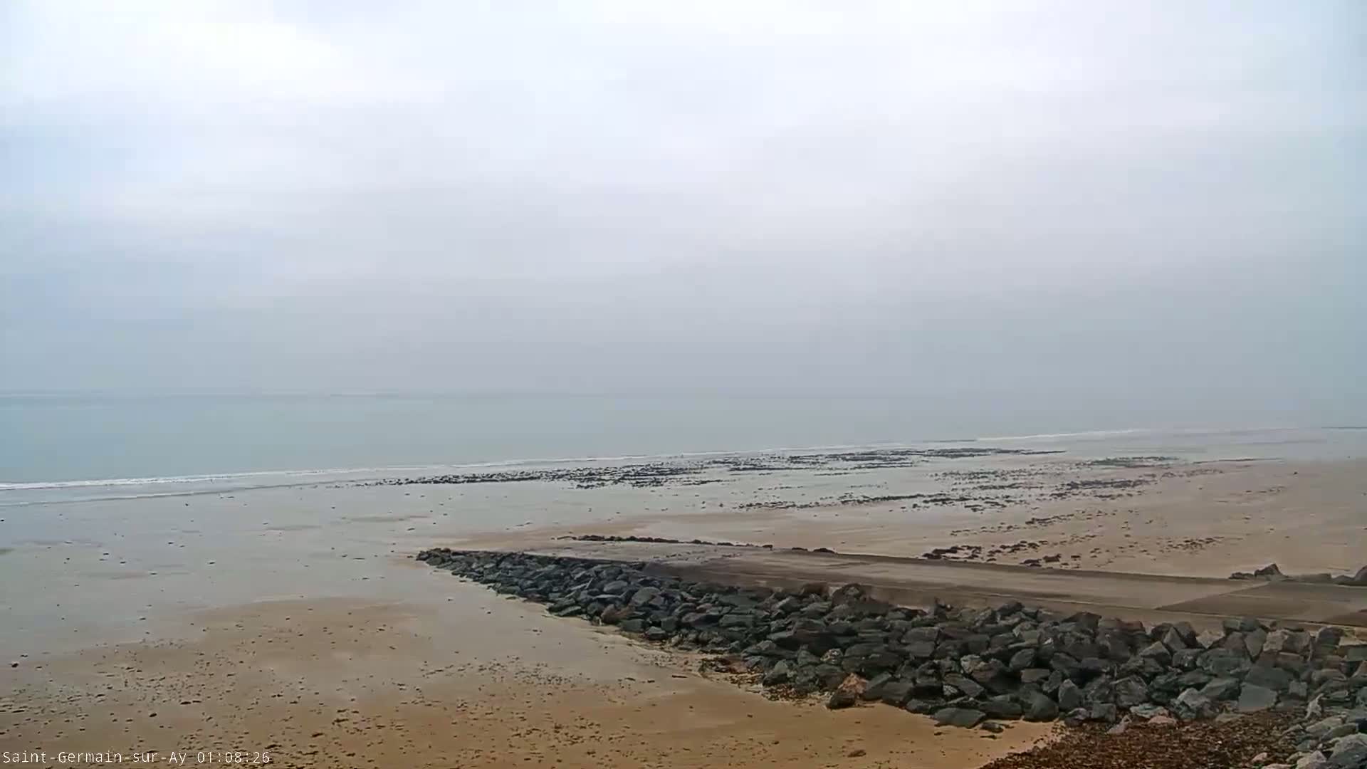 A rocky causeway extends from a sandy beach toward a calm, hazy ocean under a cloudy sky.