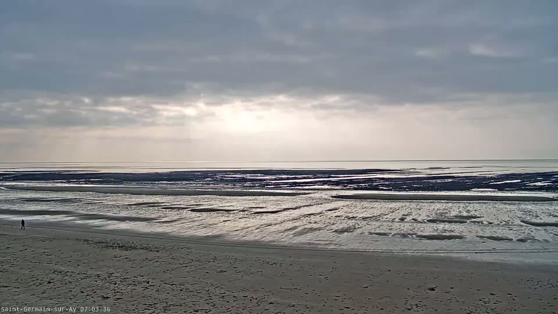 A person walks on a wide, sandy beach under a cloudy sky, with the ocean receding to reveal a vast expanse of wet sand and mudflats at low tide.