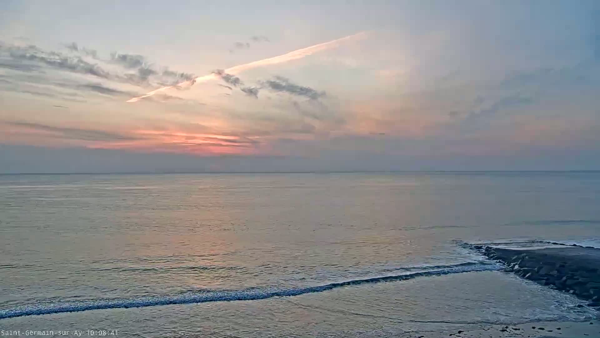A calm ocean with small waves rolls onto a rocky shore under a pastel sunset sky with scattered clouds.