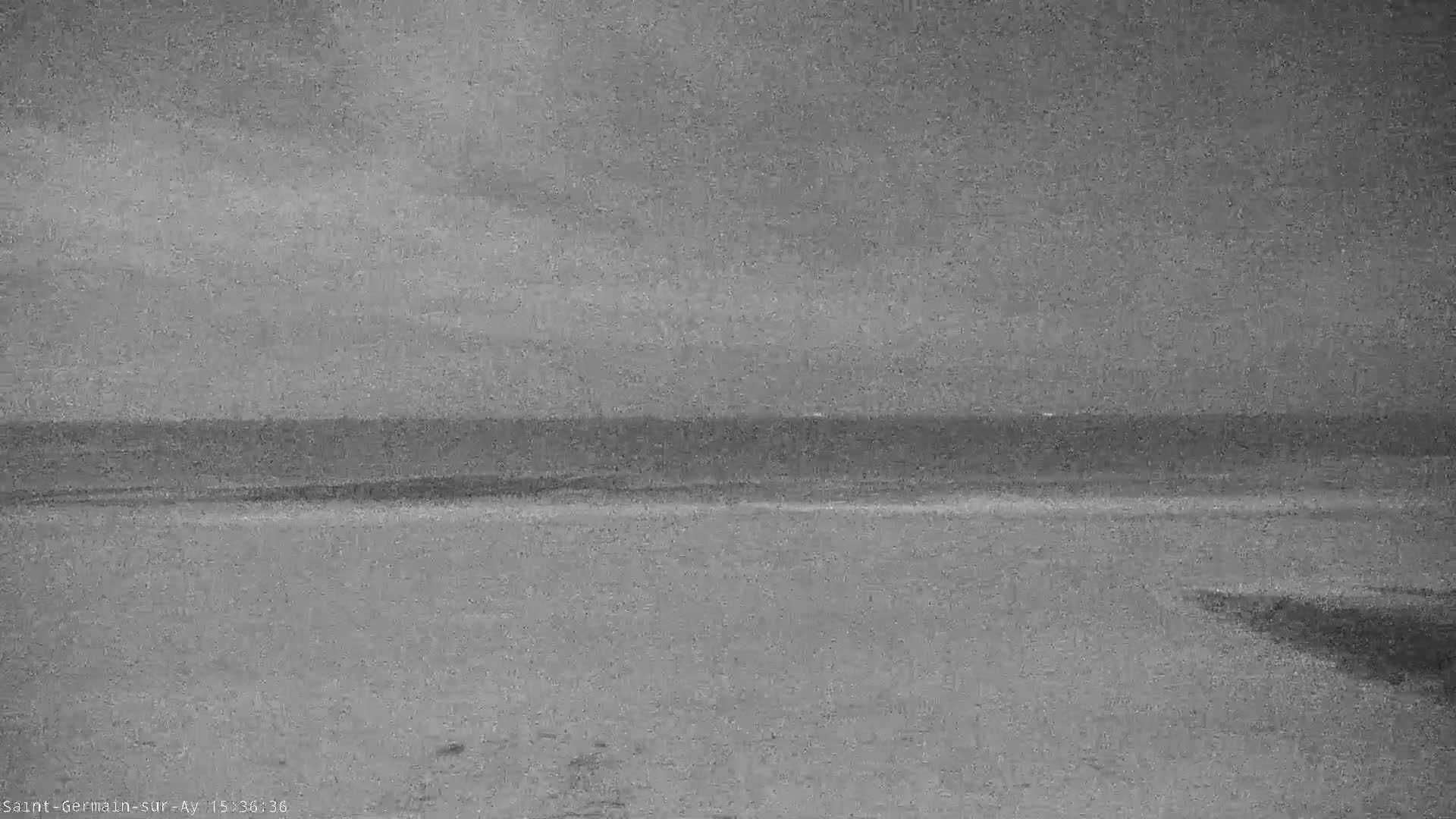 A grayscale image shows a calm ocean meeting a flat, sandy beach under an overcast sky.