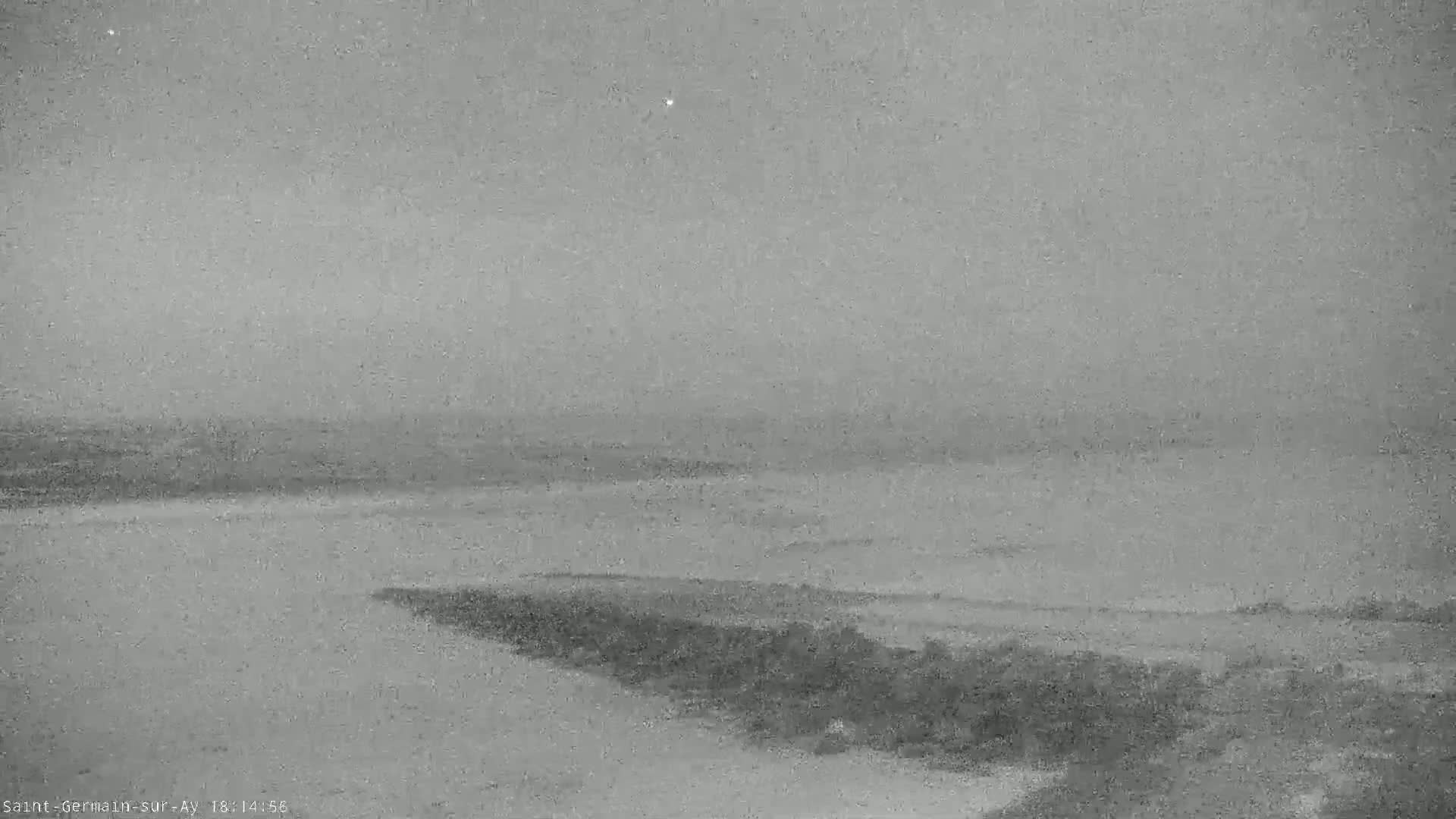 A low-quality, grayscale image shows a body of water under an overcast sky, with dark vegetation visible in the bottom right corner.