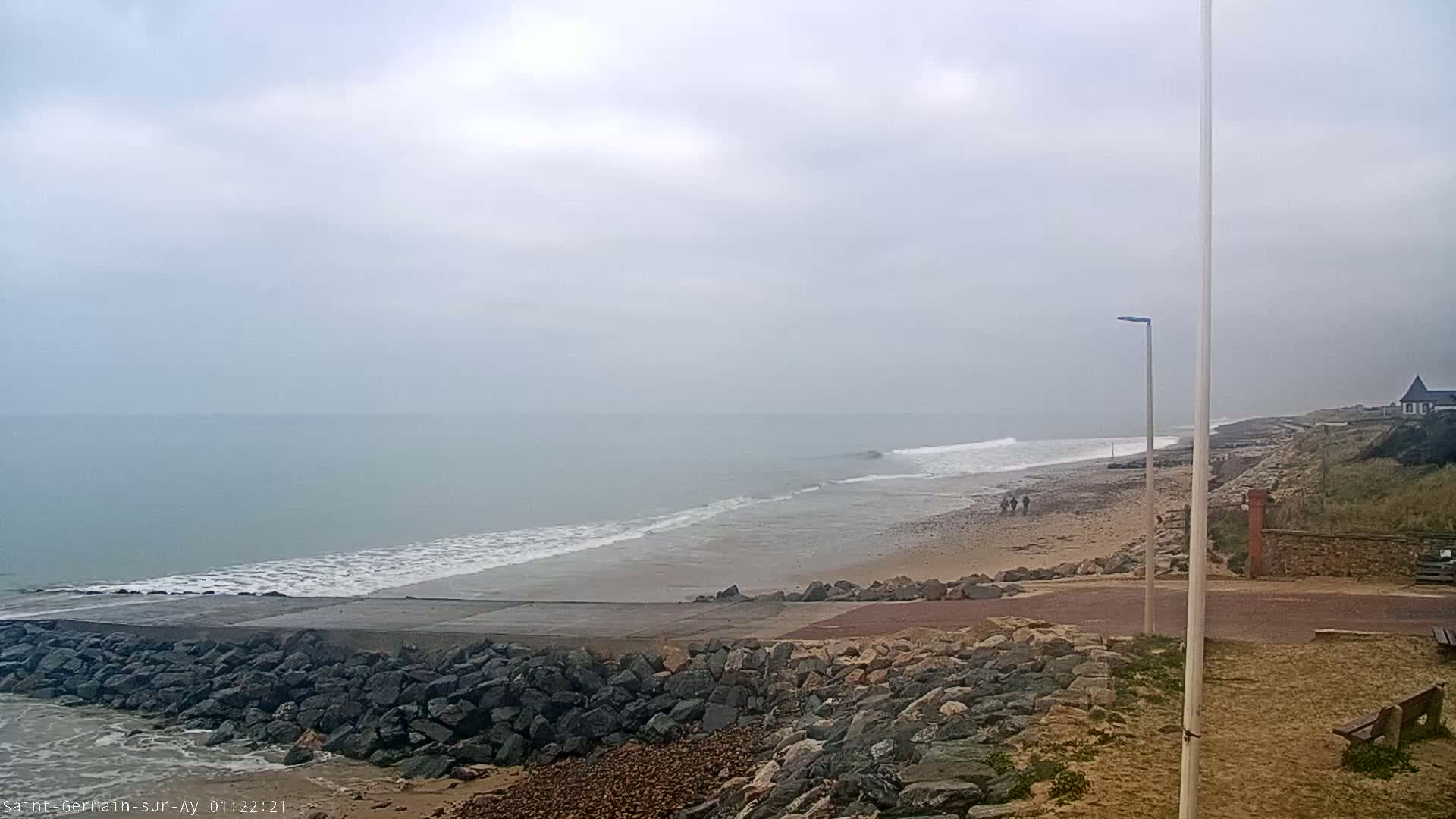 A gray, overcast day reveals a sandy beach with gentle waves lapping the shore, a rocky embankment, and a few people walking in the distance.