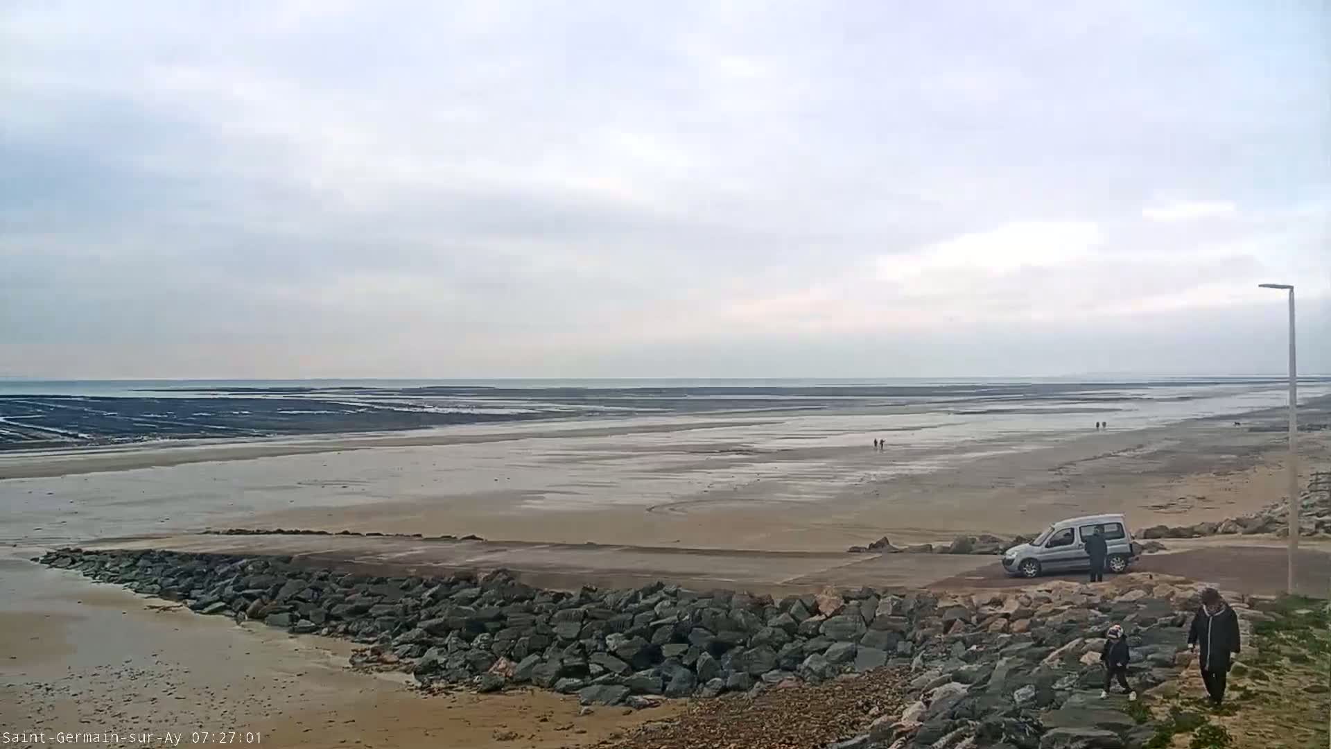 A wide, flat expanse of sand and shallow water stretches to a calm ocean under an overcast sky, with a few people and a single van visible near a rocky embankment.