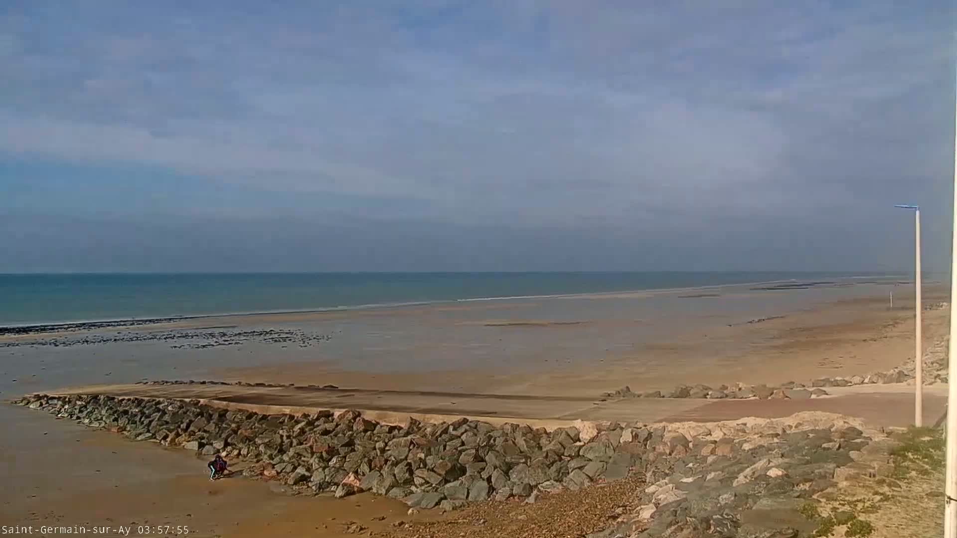 A rocky barrier protects a wide, sandy beach that stretches to a calm, light-blue ocean under a mostly clear sky.