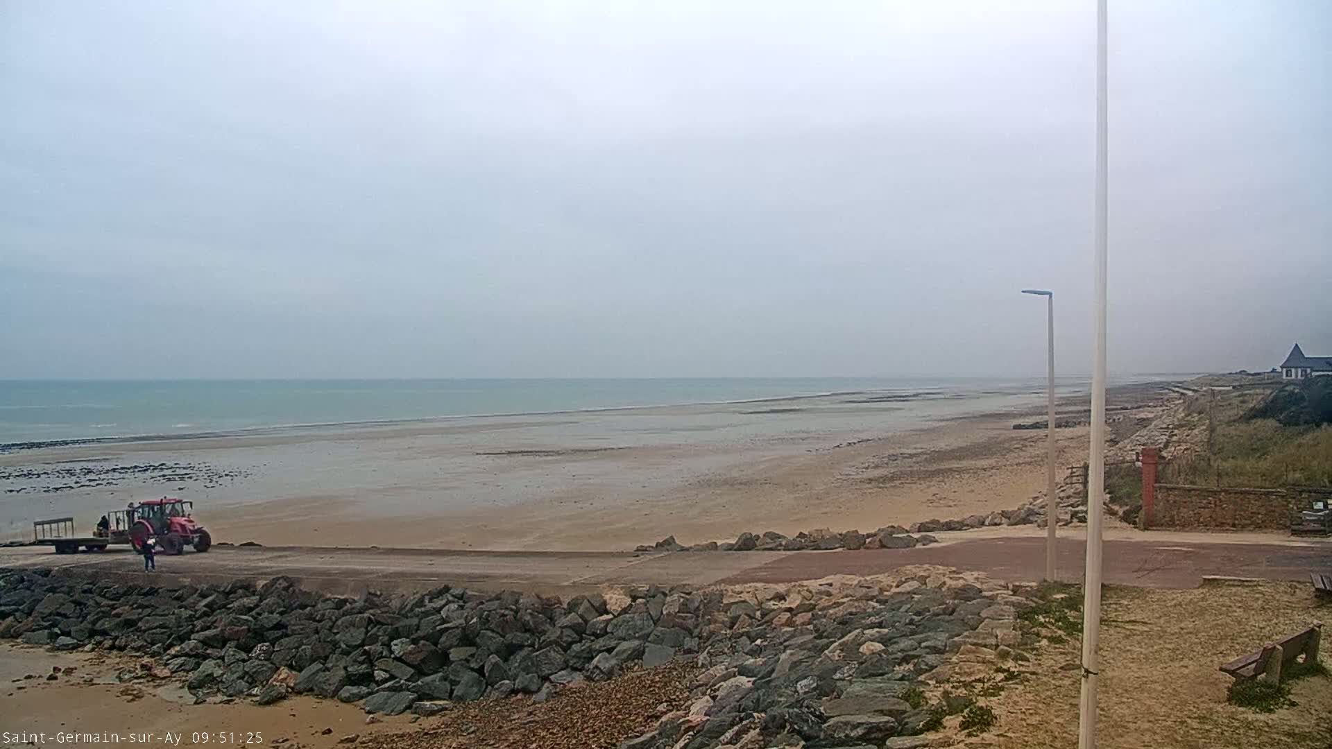A tractor and trailer are parked on a wide, sandy beach next to a rocky embankment under an overcast sky.