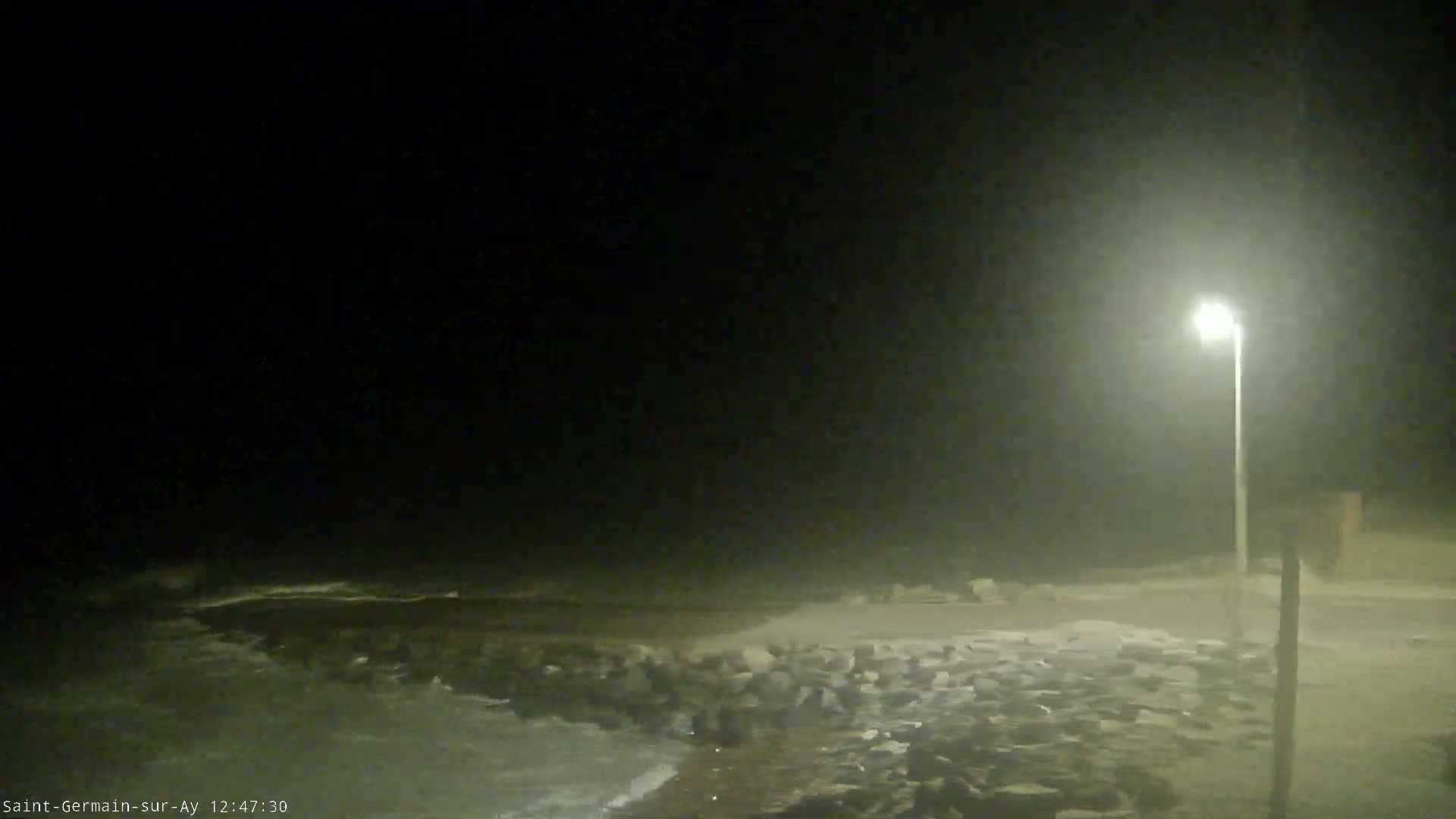 A nighttime coastal scene shows dark, misty conditions with waves lapping a rocky shore, illuminated by a single lamppost.