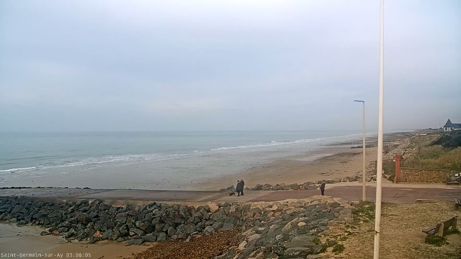 A wide sandy beach stretches along a calm, grey sea under an overcast sky, with a few people walking near a rocky embankment.