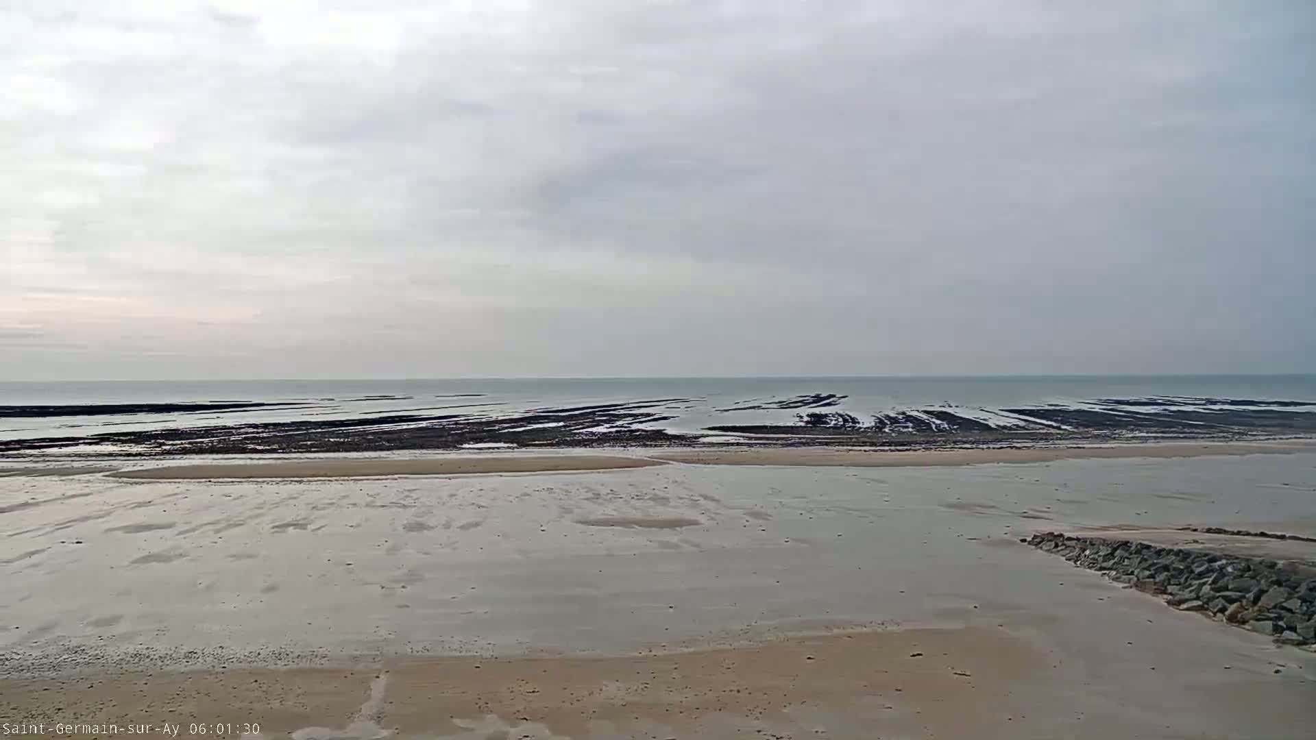A sandy beach stretches towards a calm sea under an overcast sky, revealing dark, textured rock formations at low tide.