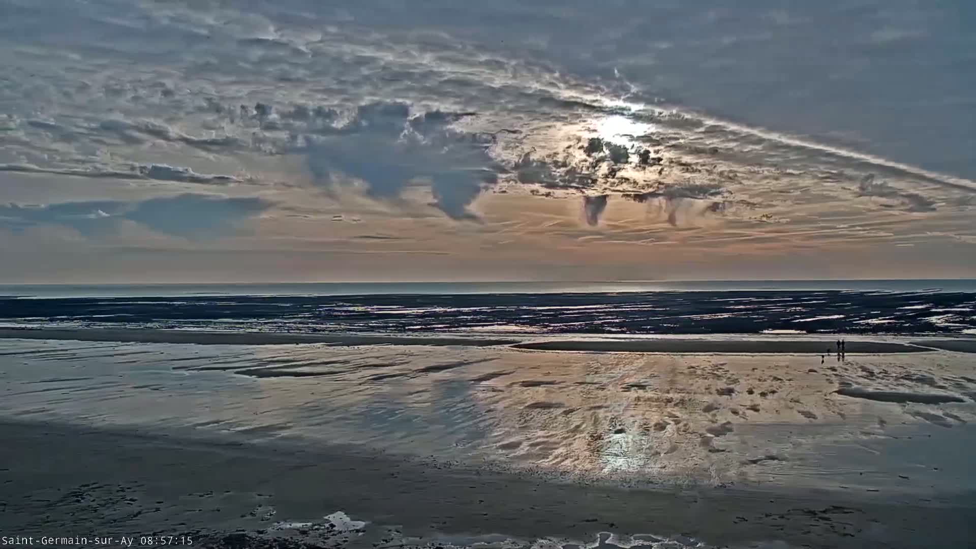 A wide expanse of wet sand reflects the sun shining through a partly cloudy sky over a calm ocean.
