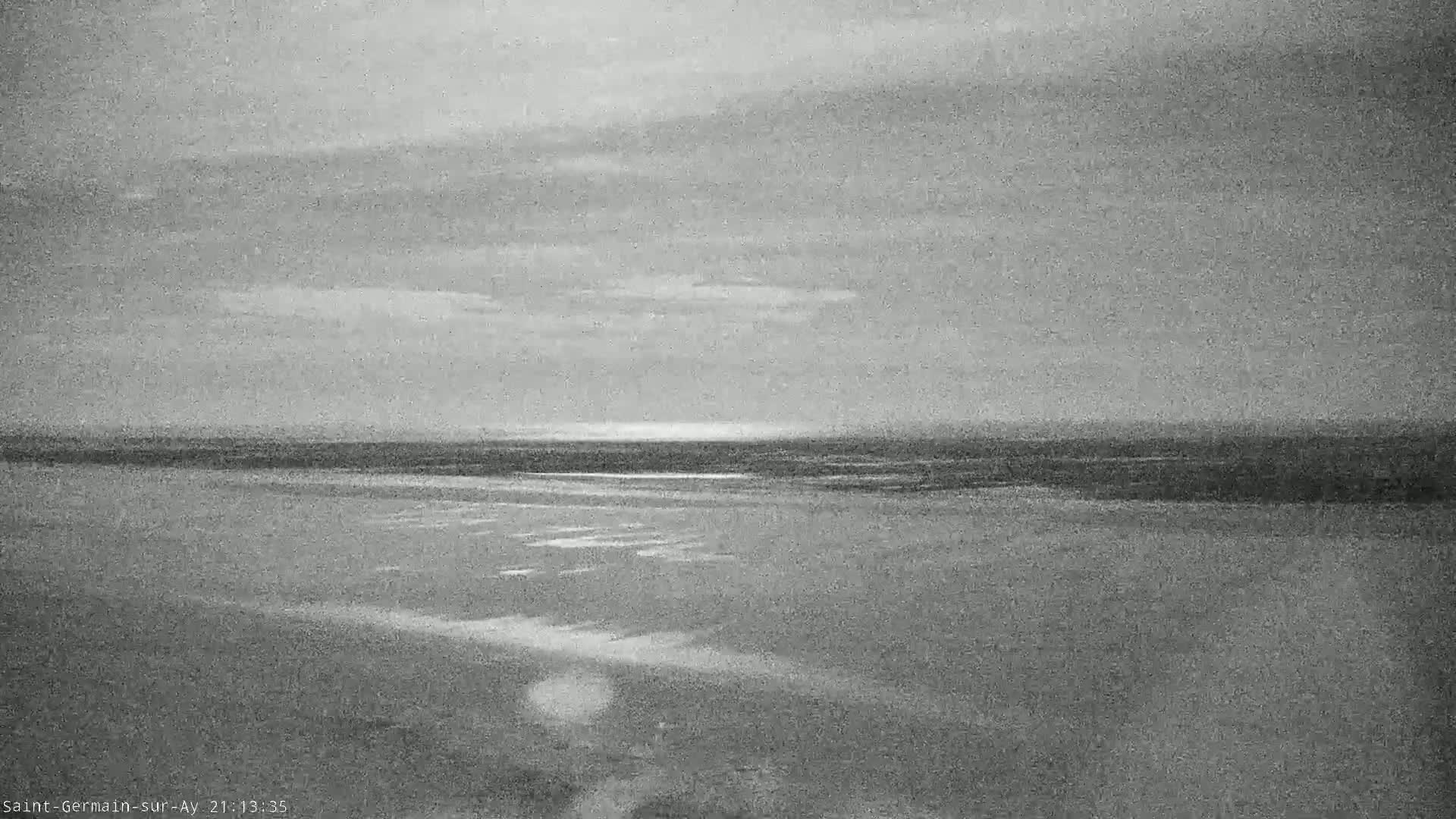 A grayscale image shows a mostly overcast sky over a flat, expansive beach and calm ocean under a pale, hazy sky.
