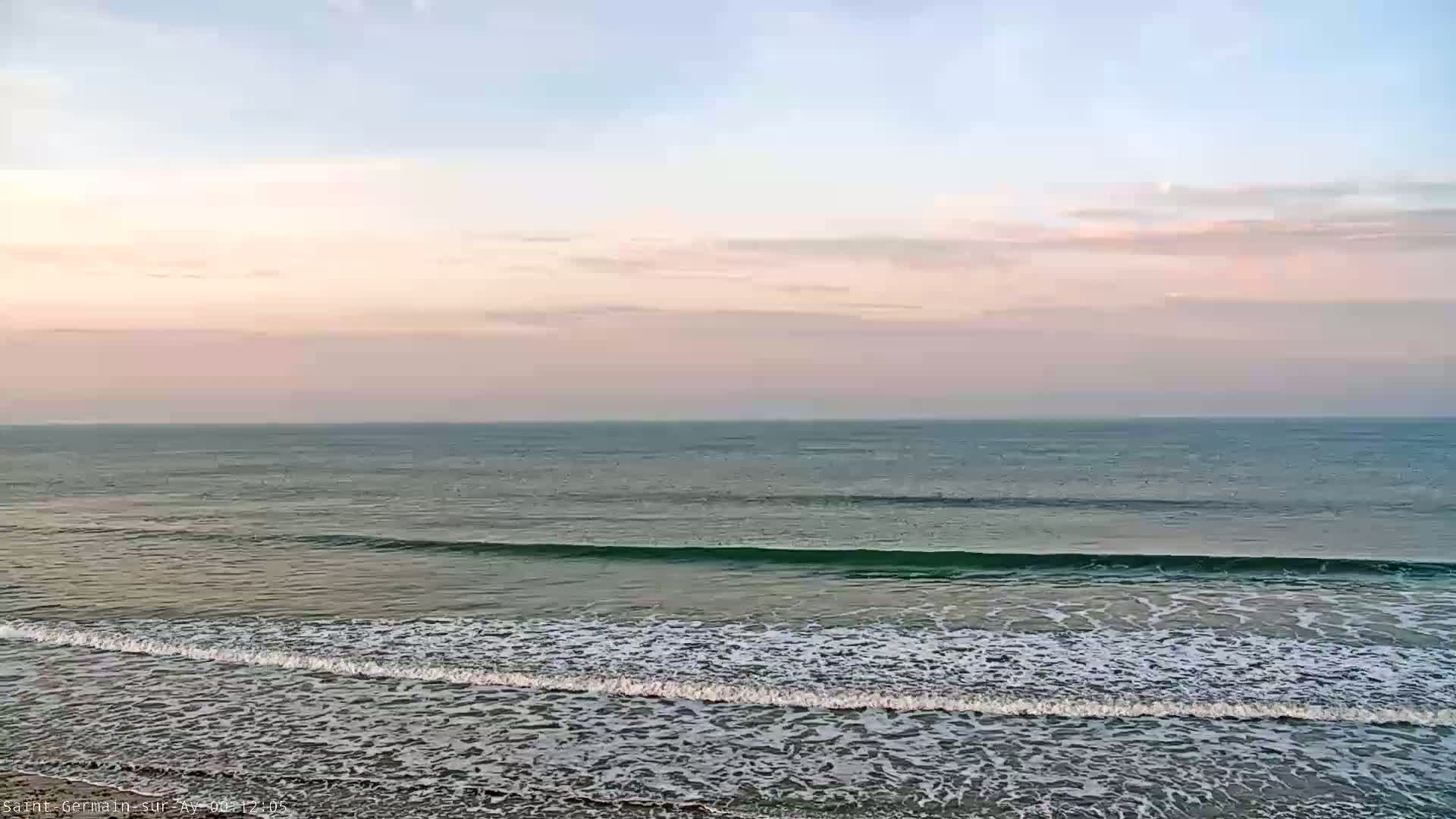 Gentle waves roll onto a beach under a pale pink and blue sky.
