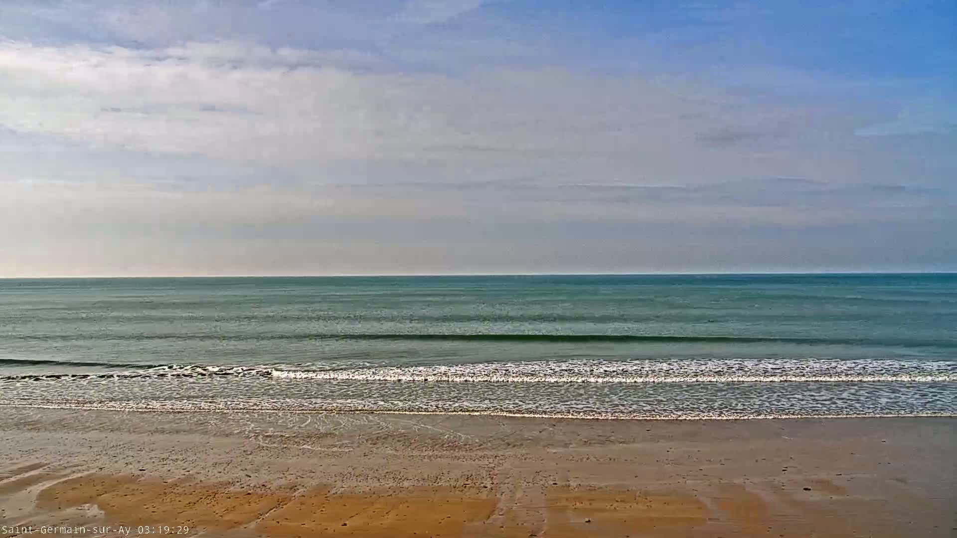 A mostly cloudy sky overlooks a calm ocean with small waves lapping a sandy beach.