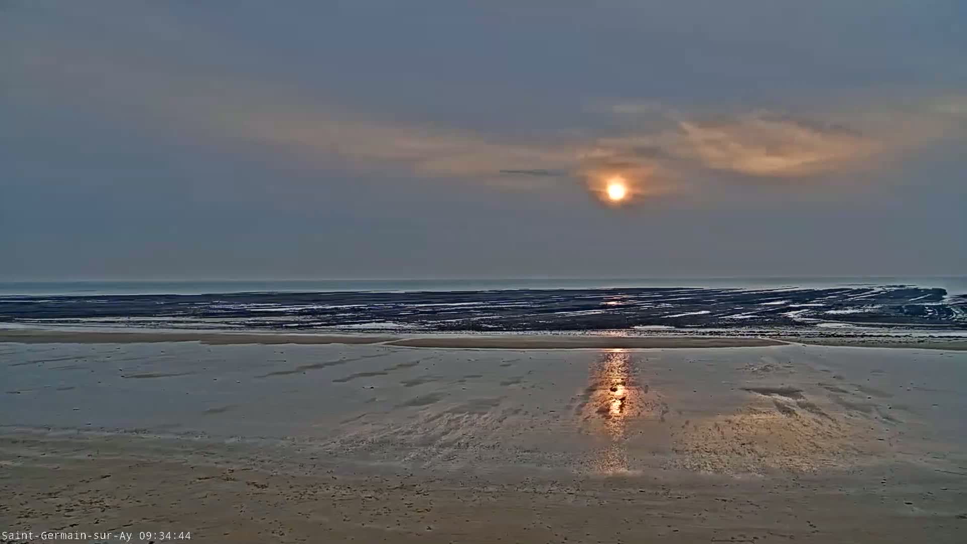 A hazy sunset over a beach with exposed, dark intertidal zones and calm, reflective water.