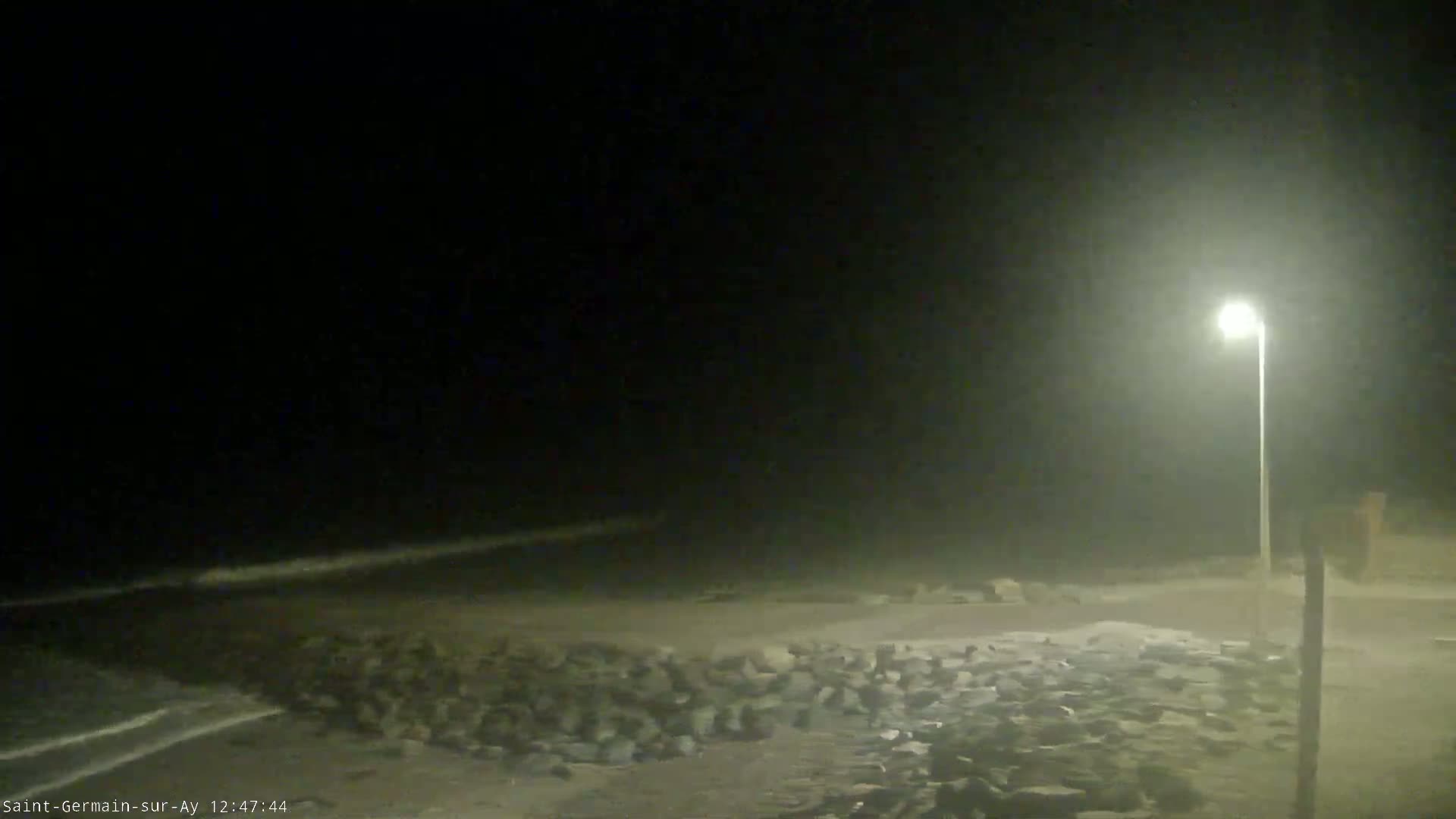 A nighttime coastal scene shows a dimly lit beach with rocks and a single lamppost under hazy or foggy conditions.